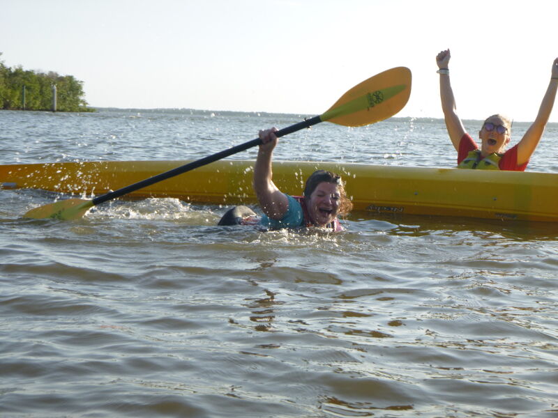 The image shows two people in the water near a yellow kayak. One person is holding a paddle and appears to be swimming or struggling in the water. The other person is in the kayak with their arms raised in the air, seemingly celebrating or cheering. The water is choppy, and there are trees visible in the background, suggesting a lake or coastal environment.